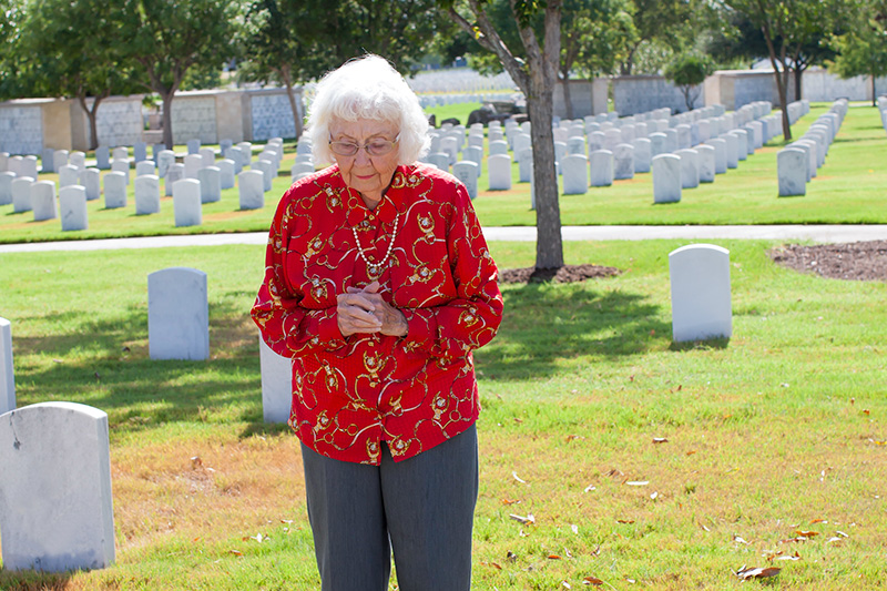 woman-standing-beside-gravestone Elderly woman standing at a cemetery, hands clasped in reflection among rows of gravestones, symbolizing remembrance and connection to loved ones.