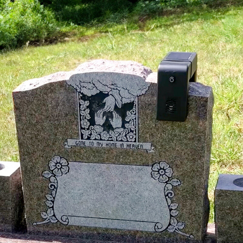 FlashBack audio memorial device securely mounted on the side of a granite headstone engraved with floral artwork and the phrase “Gone to my home in heaven,” in a grassy cemetery.