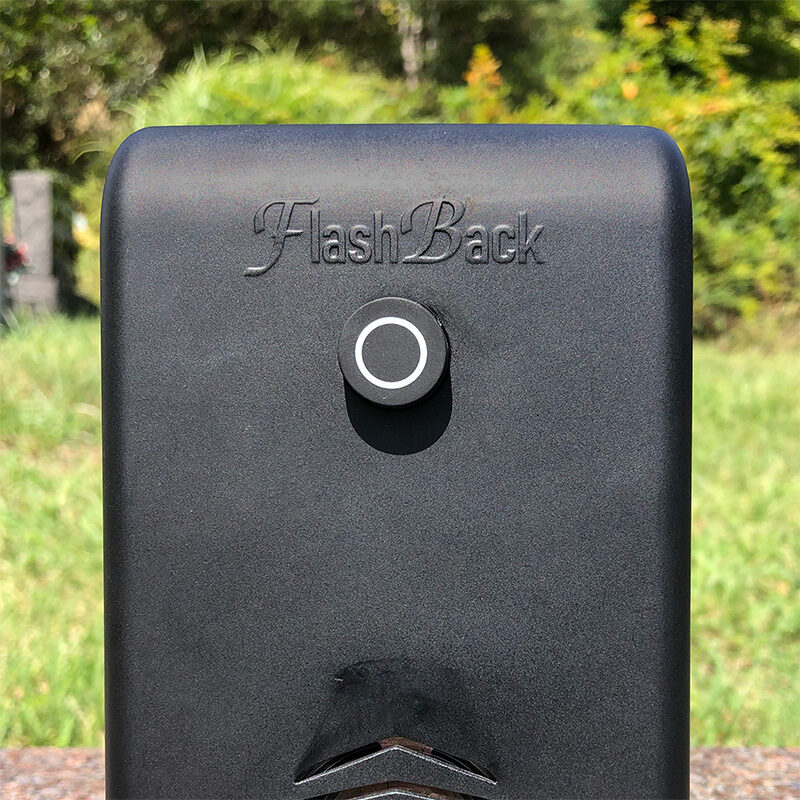 Front view of the FlashBack graveside audio device showing the engraved logo, circular play button, and speaker grille, photographed outdoors on a sunny day.