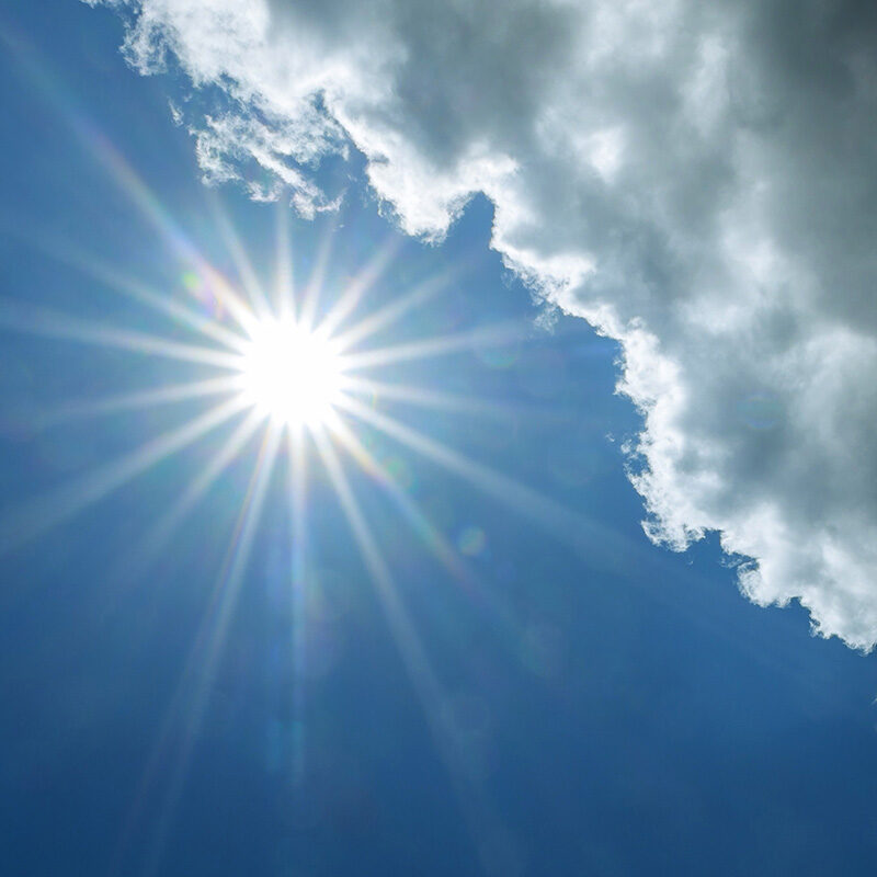 Bright sun with lens flare shining in a clear blue sky, partially obscured by the edge of a white cumulus cloud.